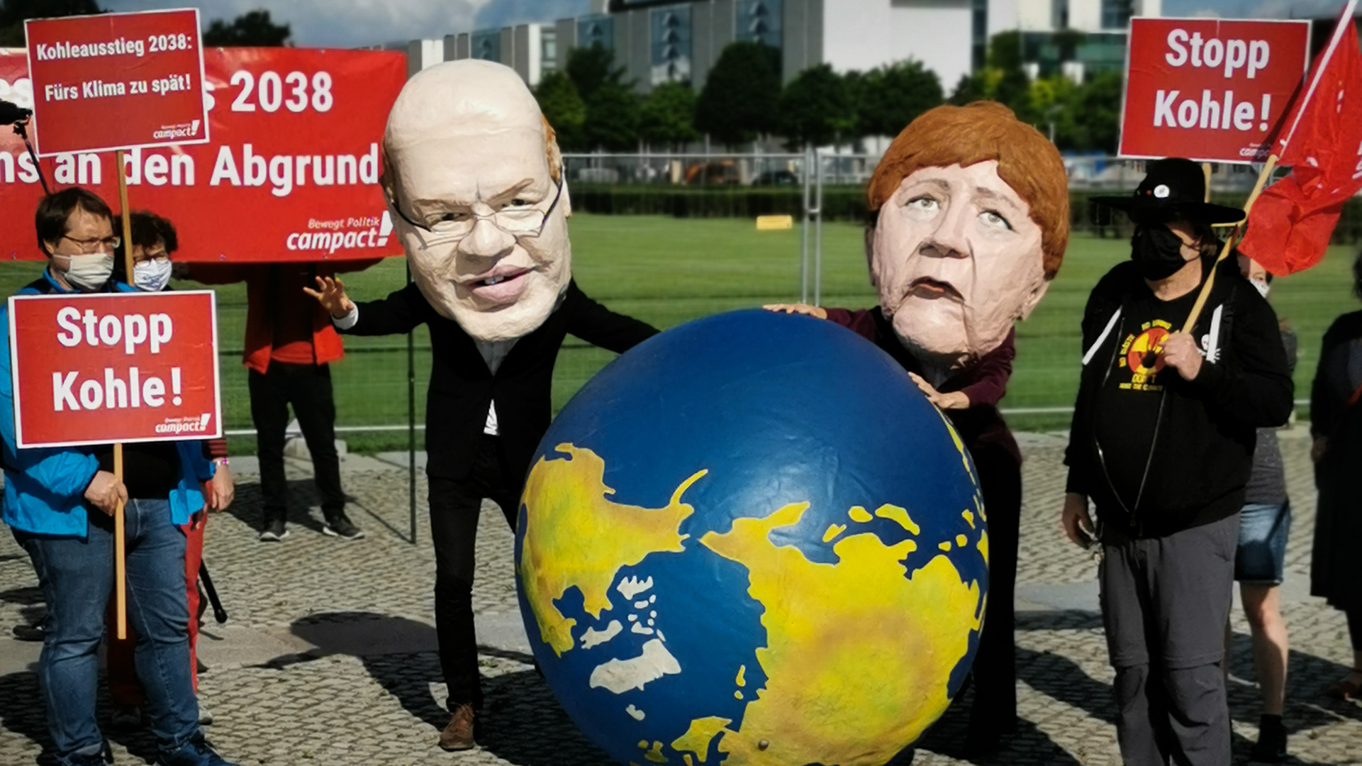 Zwei Menschen stehen als Peter Altmaier und Angela Merkelt verkleidet mit einem aufblasbaren Globus auf einer Demonstration.