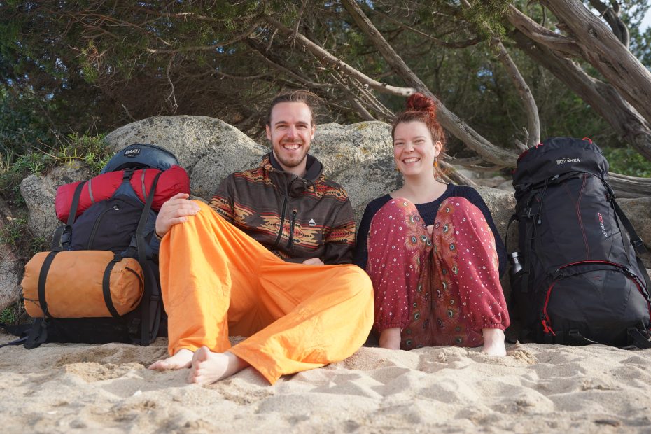 Janna und Nikolas sitzen mit ihren Backpacks auf einem sardischen Sandstrand.
