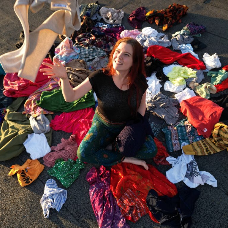 Janna sitzt in einem Berg von Kleidung und wirft grinsend ein Kleidungsstück hoch.